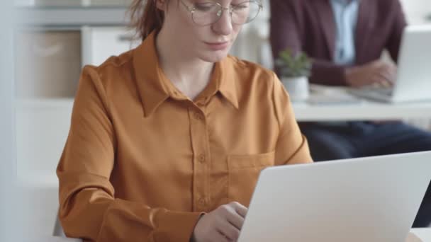 Наклонный снимок грудной клетки сфокусированной молодой белой сотрудницы в блузке и очках, сидящей за столом в офисе и быстро печатающей на ноутбуке, и мужчины среднего возраста, работающего в размытом фоне - Кадры, видео
