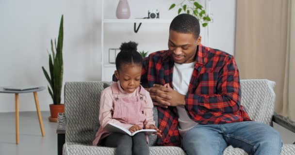 Afroamerikanischer Vater sitzt mit seiner Tochter im Wohnzimmer und hört zu, wie ein Kind während der Blockade und Quarantäne zu Hause Märchen liest, um zu lernen, junger Vater hilft dem Baby beim Lernen - Filmmaterial, Video