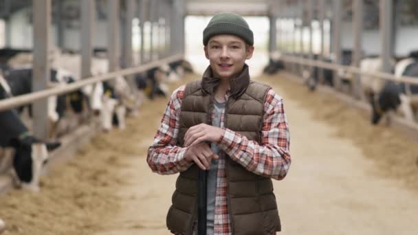 Portrait shot of cheerful 15-year-old boy in hat and puffy vest smiling for camera inside dairy farm with cows in feedlots - Footage, Video