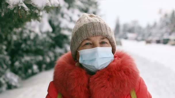 Femme âgée adulte en masque médical de protection regardant la caméra dans le parc d'hiver. Distance sociale pendant la pandémie de Coronavirus Covid 19 - Séquence, vidéo