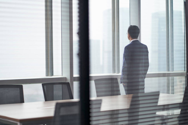 οπίσθια όψη ενός Ασιάτη επιχειρηματία που στέκεται μπροστά από το παράθυρο του γραφείου κοιτάζοντας έξω και σκεπτόμενος χέρια στην τσέπη - Φωτογραφία, εικόνα
