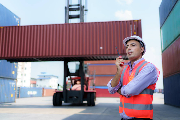 Caucasian foreman man is using walkie talkie to command his workers with crane lifting container in background in container depot terminal. - Photo, Image