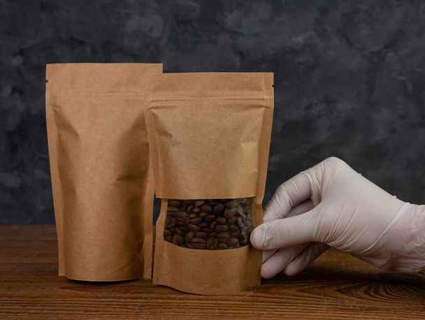 Makieta torby. Ręka w rękawicach ochronnych posiada brązowy papier kraft opakowanie z ziaren kawy drewniane tło. Opakowanie z zapięciem szablon dostawy żywności herbata pozostawia produkt wagi. Projekt reklamy sklepowej - Zdjęcie, obraz