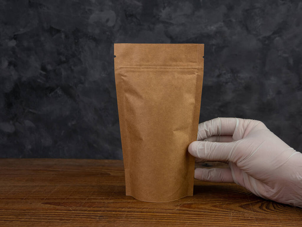 Pouch bag mockup. Hand in protective gloves holds brown kraft paper pack with coffee beans wooden background. Packaging with clasp template food delivery tea leaves weight product. Shop sale ad design - Photo, Image