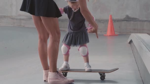 Vista posterior de una joven mujer delgada que usa ropa de calle cogida de la mano de su hija pequeña que usa coloridos cascos de seguridad, rodilleras y coderas y le enseña a patinar en el parque de skate - Imágenes, Vídeo