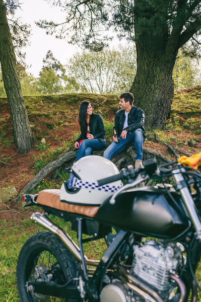 Молода пара відпочиває від поїздки на мотоциклі з пивом на відкритому повітрі з мотоциклом на передньому плані. Вибірковий фокус на парі у фоновому режимі
 - Фото, зображення