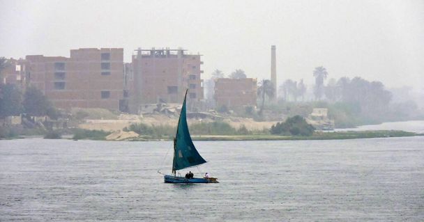 Un boutre avec une voile bleue au-dessus du Nil. Un boutre est un voilier traditionnel en bois. Au loin se trouve le contour vague d'une ville égyptienne visible - Photo, image
