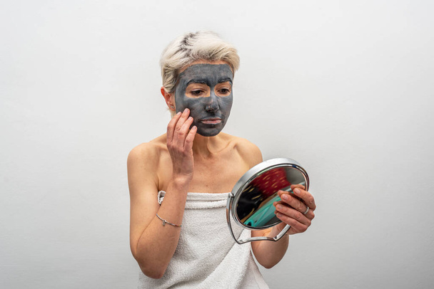 Middle aged female with clay mask looking at mirror while sitting on bed during skin care routine at home - Photo, Image