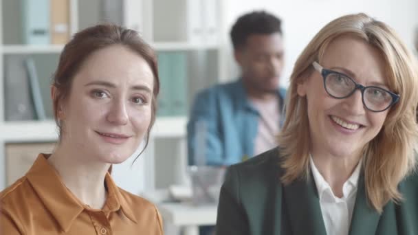 Klatka piersiowa młodej białej kobiety z rudymi włosami, w bluzce siedzącej razem z kobietą w średnim wieku w okularach i kurtce w ruchliwym biurze, patrzącej w kamerę i uśmiechniętej - Materiał filmowy, wideo