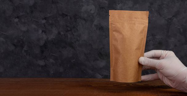 Pussi laukku mallinnus. Käsi suojakäsineet omistaa ruskea voimapaperi pakkaus kahvipapuja puinen tausta. Pakkaus malli elintarvikkeiden toimitus teetä lähtee paino tuote. Myymälä myynti mainos web bannerin suunnittelu - Valokuva, kuva
