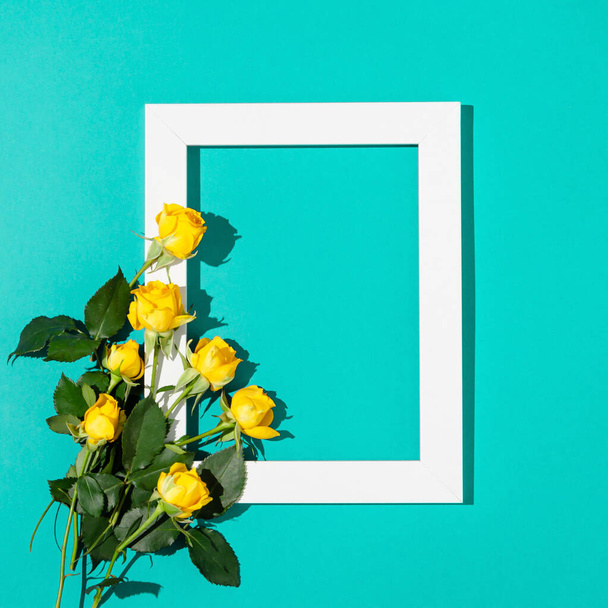 Creatief minimaal concept gemaakt van gele rozen met groene bladeren en wit frame op turquoise achtergrond. Plat gelegd. Kopieerruimte. - Foto, afbeelding