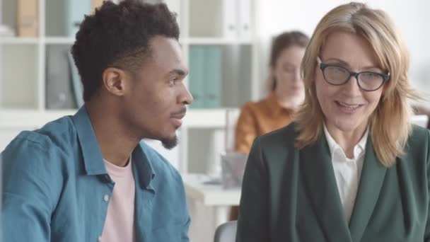 Borstfoto van een blanke vrouwelijke HR-manager van middelbare leeftijd die naast een jonge Afro-Amerikaanse stagiair zit te praten, vervolgens naar de camera kijkt en glimlacht - Video