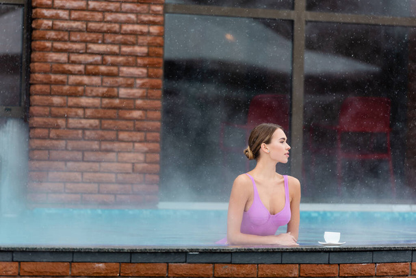 жінка в купальнику дивиться на відкритий басейн біля чашки кави на кам'яній поверхні
 - Фото, зображення