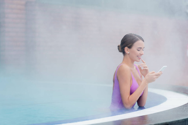 femme heureuse en maillot de bain tenant smartphone tout en se baignant dans la piscine extérieure dans le centre de spa  - Photo, image