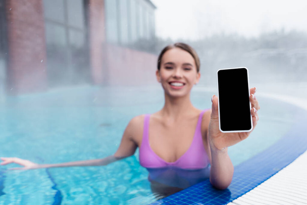 mujer borrosa y alegre sosteniendo teléfono inteligente con pantalla en blanco mientras se baña en la piscina al aire libre - Foto, imagen