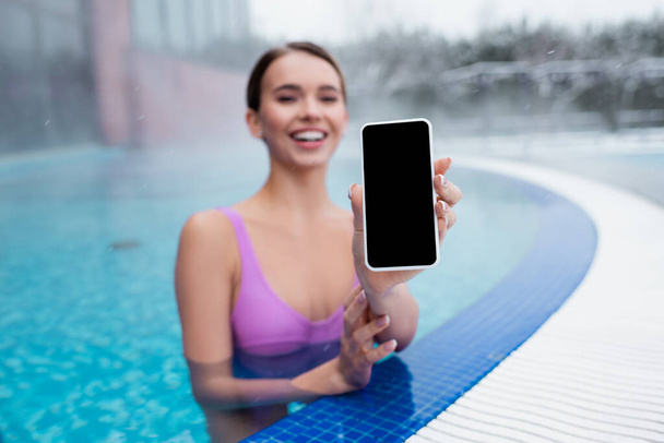 mujer alegre sosteniendo teléfono inteligente con pantalla en blanco mientras se baña en la piscina al aire libre - Foto, imagen
