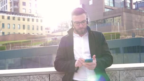 Hidasliikkeinen nuori valkoihoinen parrakas mies ulkona kuuntelee musiikkia kuulokkeet ja messaging älypuhelimen kädensija hymyilevä positiivinen ja rento - Materiaali, video