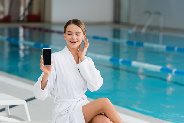 mujer alegre en albornoz blanco sosteniendo teléfono inteligente con pantalla en blanco en el centro de spa  - Foto, Imagen