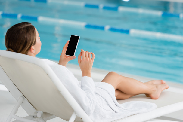 mujer joven descansando en la silla de cubierta y el uso de teléfono inteligente con pantalla en blanco en el centro de spa  - Foto, Imagen