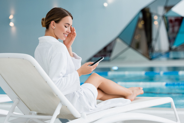 happy woman resting on deck chair and holding smartphone near pool - Foto, afbeelding