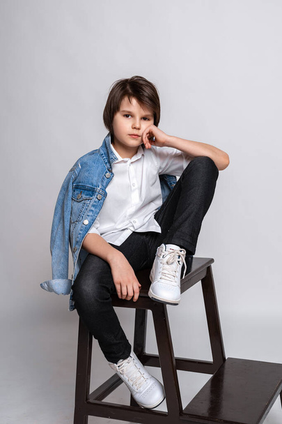 Hermoso niño de la moda 11-13 años de edad se sienta en una silla en el estudio. Vestido con camisa blanca, jeans negros, chaqueta jean y zapatillas blancas. Prueba de modelo. Concepto moda y gente - Foto, Imagen