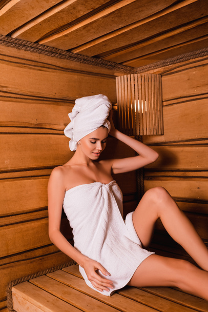 Mujer joven envuelta en toallas sentada en el asiento en la sauna de madera  - Foto, Imagen