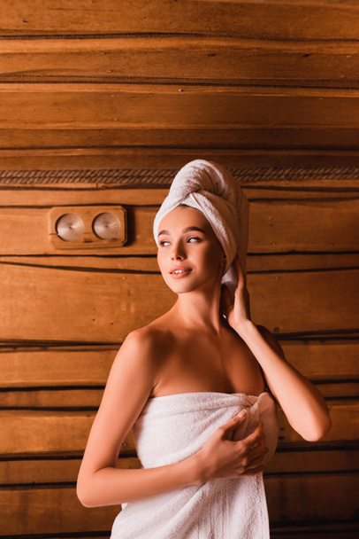 Mujer sonriente en toallas blancas mirando hacia otro lado en sauna de madera  - Foto, Imagen