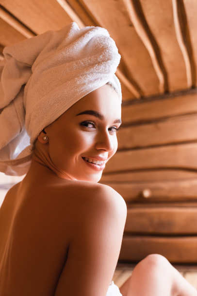 vrouw met handdoek op hoofd glimlachen op camera in sauna op wazig achtergrond  - Foto, afbeelding