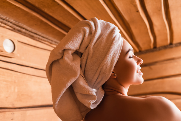 улыбающаяся женщина с полотенцем на голове и обнаженными плечами отдыхает в сауне  - Фото, изображение