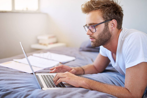 Hezký mladý muž v pyžamu nosí brýle a pomocí notebooku při ležení v posteli v dopoledních hodinách - Fotografie, Obrázek