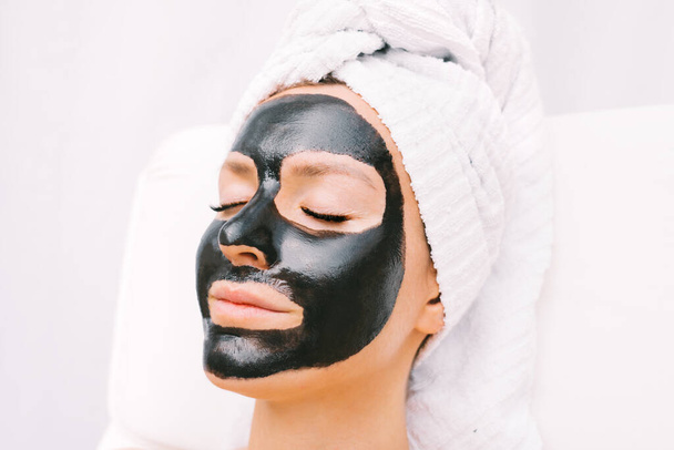 Красива жінка з рушником на голові розслабляється чорною очищаючою маскою для обличчя. Косметологія та скінарій. Красиві процедури. Спа-терапія
. - Фото, зображення