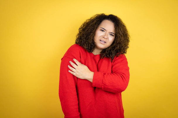 Junge Afroamerikanerin trägt roten Pullover über gelbem Hintergrund mit Schmerzen an der Schulter und einem schmerzhaften Gesichtsausdruck - Foto, Bild