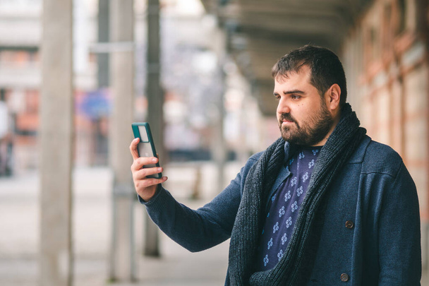 Bärtiger Mann im blauen Pullover steht mit Smartphone auf der Straße. - Foto, Bild