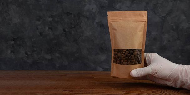 ポーチバッグモックアップ。保護手袋の手は、コーヒー豆の木の背景と茶色のクラフト紙パックを保持しています。包装テンプレート食品配達お茶は重量製品を残します。ショップセール広告ウェブバナーデザイン - 写真・画像
