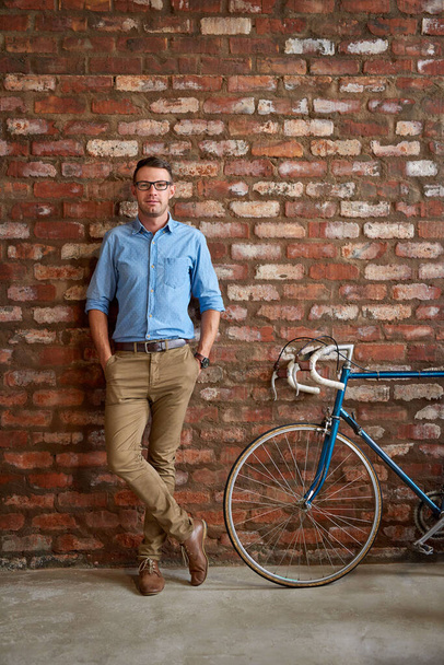 Retrato de un joven hombre de negocios vestido casualmente de pie junto a una bicicleta apoyada contra una pared de ladrillo en una oficina - Foto, Imagen