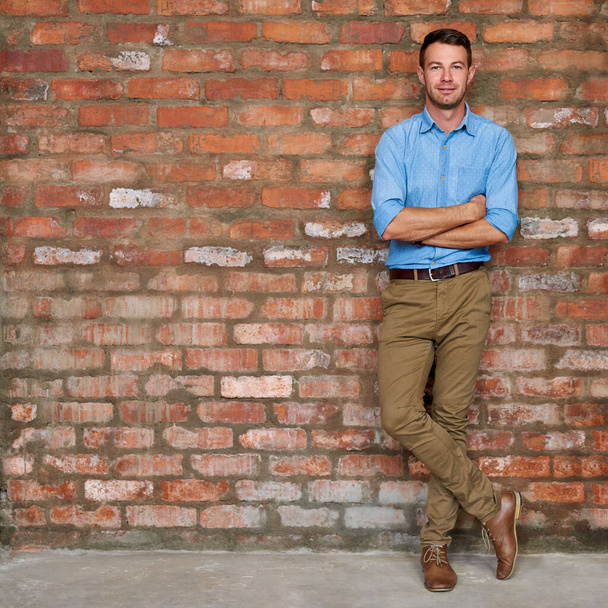 Portrait d'un jeune homme d'affaires vêtu avec désinvolture, les bras croisés contre un mur de briques dans un bureau - Photo, image