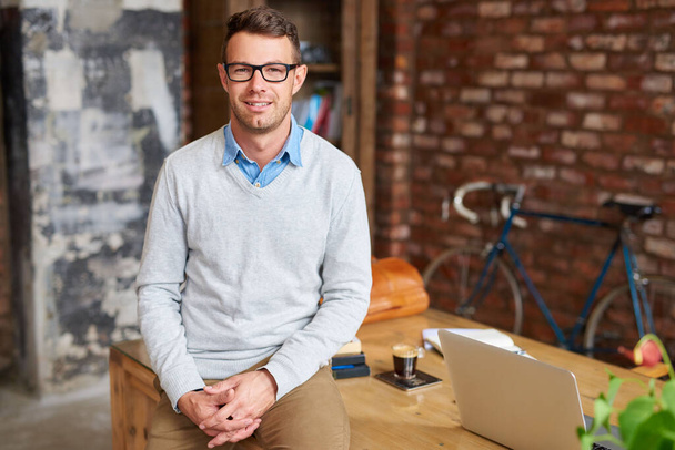 Portrait of a casually dressed young businessman wearing glasses and sitting on a desk in a trendy brick walled office - Photo, Image