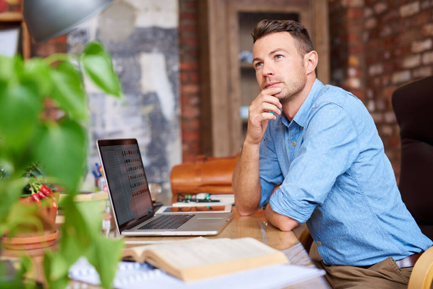 Jeune homme d'affaires habillé de façon décontractée réfléchissant tout en travaillant sur un ordinateur portable à un bureau dans un bureau   - Photo, image