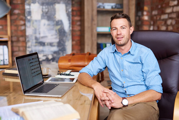 Portret van een casual geklede jonge zakenman die op een laptop werkt terwijl hij achter een bureau in een kantoor zit   - Foto, afbeelding