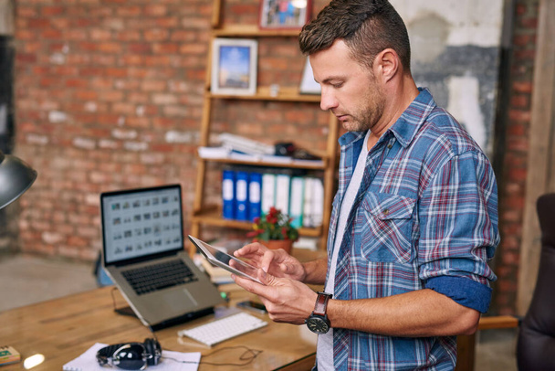 Tuğladan duvarlarla örülü bir ofiste masa başında dururken tablet kullanan sıradan giyinmiş genç bir tasarımcı.   - Fotoğraf, Görsel