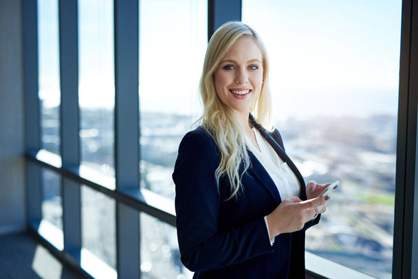 Портрет усміхненої молодої бізнес-леді, яка тримає мобільний телефон, стоячи біля вікон з видом на місто в сучасній офісній будівлі
 - Фото, зображення