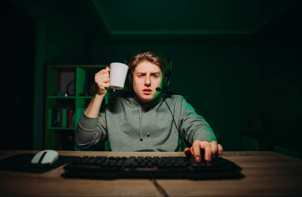 Portrait d'un joueur fatigué dans un casque avec une tasse dans ses mains en jouant à des jeux en ligne sur l'ordinateur et en regardant attentivement la caméra. - Photo, image