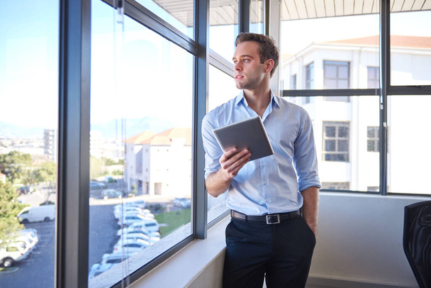 Όμορφος νεαρός επιχειρηματίας, στέκεται στο γραφείο του με το χέρι στην τσέπη του και κρατώντας μια ψηφιακή ταμπλέτα, ενώ κοιτάζει έξω από το παράθυρο του γραφείου του και σκέφτεται - Φωτογραφία, εικόνα