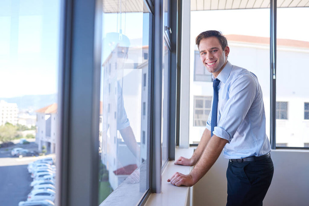 Porträt eines freundlichen jungen Geschäftsmannes, der in seinem Büro an den Fenstern steht und in die Kamera lächelt - Foto, Bild