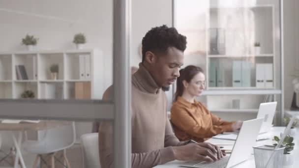 Obloukový záběr pohledného mladého afroamerického muže ve svetříku, sedícího za stolem v zaneprázdněné kanceláři, pracujícího na laptopu, a pak se s veselým a sebevědomým úsměvem obracejícího ke kameře - Záběry, video
