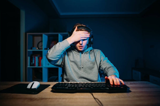 Un joueur perdant dans un casque se trouve la nuit dans une pièce avec une lumière bleue à une table à un ordinateur et fait un geste de paume du visage. Concept de jeu. - Photo, image