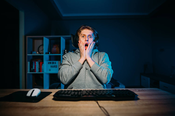 Portret van een geschokte gamer in casual kleding en een headset op zijn hoofd zitten 's nachts thuis op de computer en staren naar het scherm in angst. Jongeman stream horror spel en bang. - Foto, afbeelding