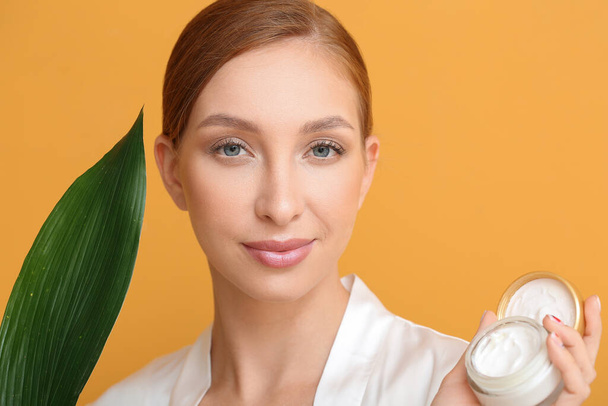 Beautiful young woman with facial cream and tropical leaf on color background, closeup - Foto, afbeelding