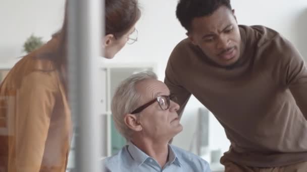 Close-up shot van gefrustreerde oudere blanke man aan het bureau, die klaagt over werkproblemen bij multinationale junior collega 's, en zwarte man die naar laptopscherm kijkt en advies geeft - Video