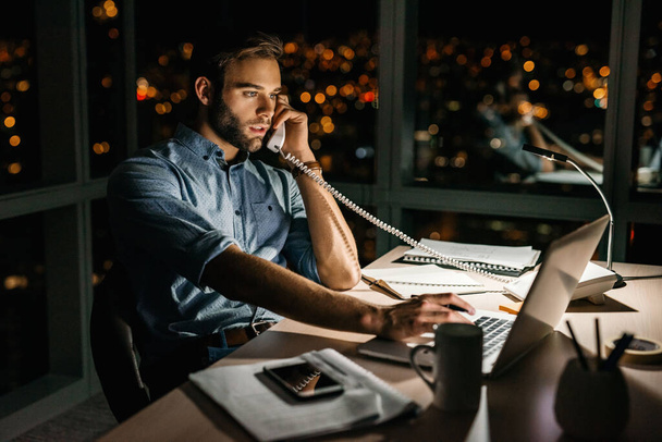 Jonge zakenman praat aan de telefoon terwijl hij overuren maakt aan zijn bureau laat in de nacht voor ramen met uitzicht op de stad  - Foto, afbeelding
